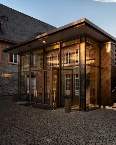 Ein modernes Gebäude mit einer Glasfassade und einer schrägen Treppe, umgeben von einem gepflasterten Platz. Die Architektur kombiniert zeitgenössische Elemente mit traditionellen Materialien.