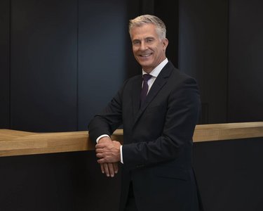 A man in a suit stands smiling at a wooden counter in a modern, dimly lit room.