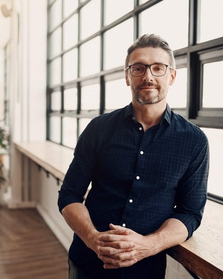 Ein Mann mit Brille und einem dunkelblauen Hemd steht an einem Tisch in einem modernen Büro mit großen Fenstern. Im Hintergrund sind Arbeitsplätze und Pflanzen sichtbar.