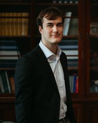 A young man in a black suit and white shirt stands in front of a bookshelf with a neutral pose.