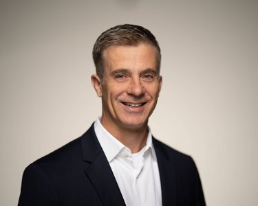 Ein lächelnder Mann mit kurzen, grauen Haaren trägt einen dunklen Anzug und ein weißes Hemd. Der Hintergrund ist neutral und hell.