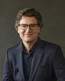 Ein Mann mit lockigem, braunem Haar und Brille trägt einen dunklen Anzug und lächelt freundlich vor einem neutralen Hintergrund.