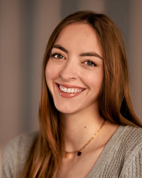 Eine lächelnde Frau mit langen, braunen Haaren trägt einen grauen Pullover und eine Halskette. Der Hintergrund ist unscharf und hell.