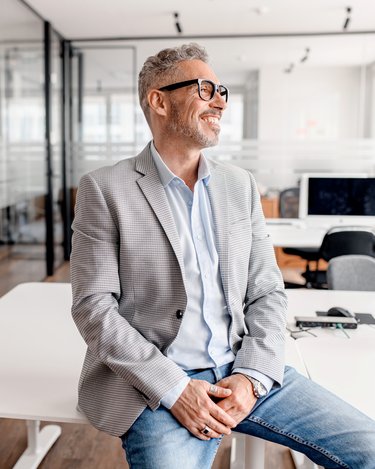 Ein Mann mit Brille sitzt auf einem Tisch in einem modernen Büro. Er trägt einen grauen Blazer und eine hellblaue Hemd. Im Hintergrund sind Computerarbeitsplätze und Glaswände sichtbar.