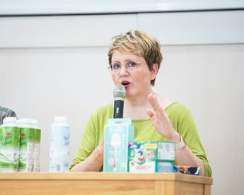 A woman with short blonde hair and glasses is speaking into a microphone. She is wearing a green top and gesturing with her hand. On the table in front of her are various drink containers.