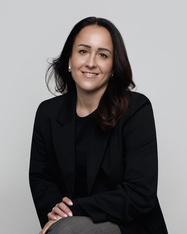 Eine Frau mit langen, dunklen Haaren sitzt in einem schwarzen Blazer und lächelt freundlich. Der Hintergrund ist neutral und hell.