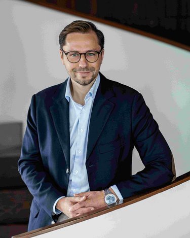 Ein Mann mit Brille steht an einer geschwungenen Treppe, trägt einen dunkelblauen Blazer und ein helles Hemd.