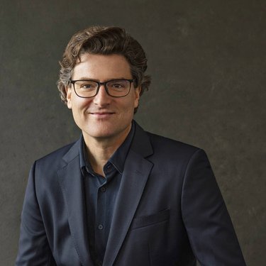 Ein Mann mit lockigem Haar und Brille trägt einen dunklen Anzug und sitzt vor einem neutralen Hintergrund.