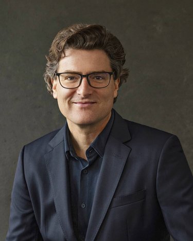 Ein Mann mit lockigem Haar und Brille trägt einen dunklen Anzug und sitzt vor einem neutralen Hintergrund.