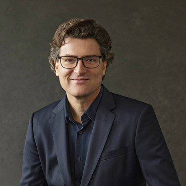Ein Mann mit lockigem Haar und Brille trägt einen dunklen Anzug und sitzt vor einem neutralen Hintergrund.