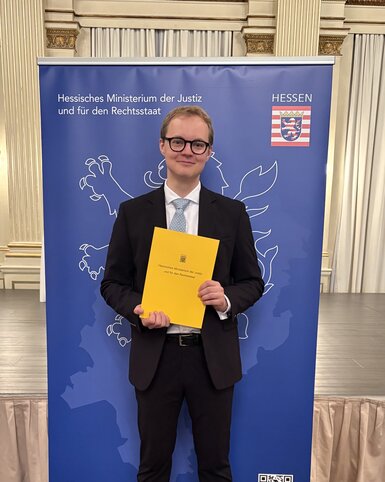 Ein junger Mann in Anzug hält ein gelbes Dokument und steht vor einem Banner des Hessischen Ministeriums der Justiz.