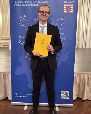 A young man in a suit is holding a yellow document and standing in front of a banner of the Hesse Ministry of Justice.