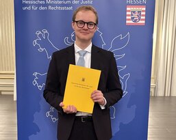 Ein junger Mann in Anzug hält ein gelbes Dokument und steht vor einem Banner des Hessischen Ministeriums der Justiz.