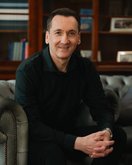 Ein Mann in einem schwarzen Hemd sitzt auf einem grauen Sofa und lächelt. Im Hintergrund sind Bücherregale sichtbar.
