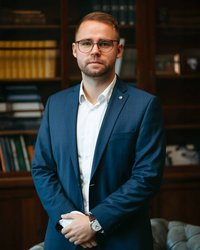 A man in a suit stands with his arms crossed in front of a bookshelf. He is wearing glasses and looking directly at the camera.