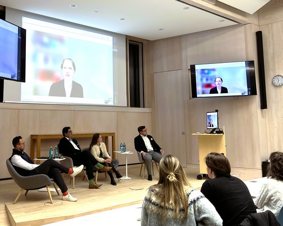 Eine Diskussionsrunde mit vier Personen, die auf einem Podium sitzen, während eine Person über einen Bildschirm zugeschaltet ist.