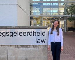 A person stands next to a sign that reads 'Regsggeleerdheid Law' in front of a modern building.