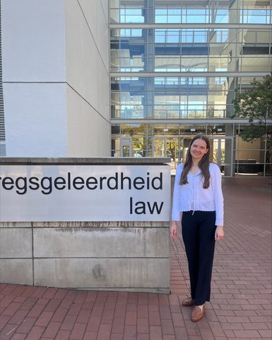 A person stands next to a sign that reads 'Regsggeleerdheid Law' in front of a modern building.