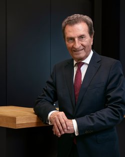 An older man in a suit stands at a wooden bar, smiling and looking into the camera. The background is dark and modern.