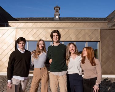 Fünf junge Erwachsene stehen lächelnd vor einem modernen Gebäude mit goldener Fassade. Sie tragen legere, stilvolle Kleidung und posieren in einer entspannten Haltung.