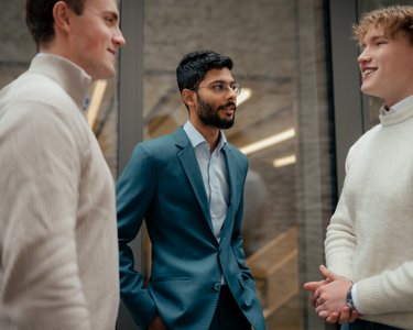 Drei Männer in formeller Kleidung unterhalten sich in einem modernen Raum. Zwei tragen helle Pullover, einer einen Anzug.