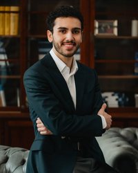 A young man in a dark suit sits on a sofa, smiling with his arms crossed, in front of a bookshelf.