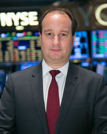 Mann in Anzug mit Krawatte steht vor einem Bildschirm mit Börsendaten. Hintergrund zeigt die NYSE.