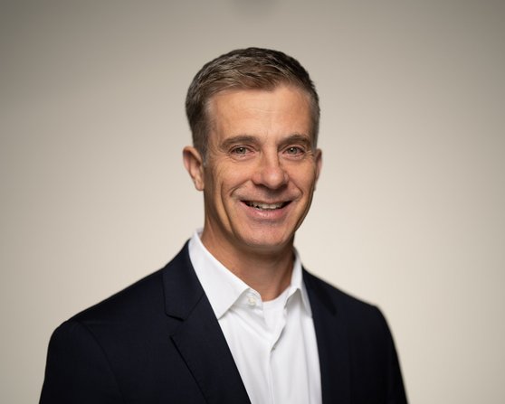 A man with short, gray hair is wearing a dark suit and a white shirt. He is smiling warmly at the camera.