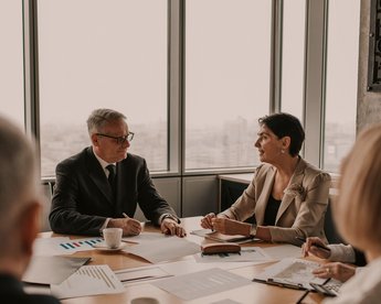 Zwei Personen in formeller Kleidung sitzen an einem Konferenztisch und diskutieren. Auf dem Tisch liegen Dokumente und ein Kaffeebecher. Im Hintergrund sind große Fenster mit Blick auf eine Stadtlandschaft.