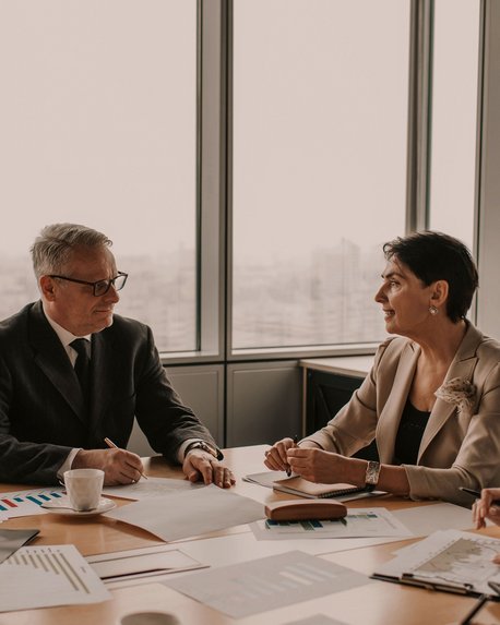 Zwei Personen in formeller Kleidung sitzen an einem Konferenztisch und diskutieren. Auf dem Tisch liegen Dokumente und ein Kaffeebecher. Im Hintergrund sind große Fenster mit Blick auf eine Stadtlandschaft.
