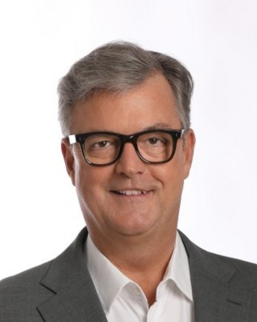 Ein Mann mit Brille und grauem Anzug lächelt in die Kamera. Der Hintergrund ist hell und neutral.