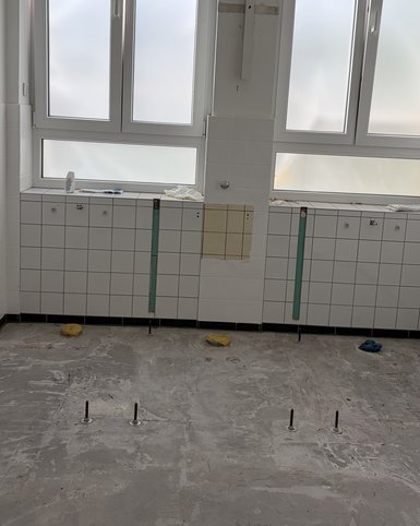 Leere, geflieste Wand mit zwei großen Fenstern und sichtbaren Installationsvorbereitungen. Der Boden ist uneben und zeigt einige Befestigungselemente.
