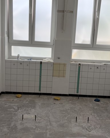 Empty, tiled wall with two large windows and visible installation preparations. The floor is uneven and shows some fasteners.
