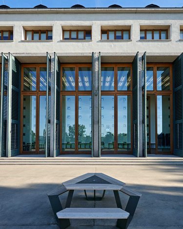 Ein modernes Gebäude mit großen Fenstern und offenen Fensterläden, die einen Blick auf den Innenraum ermöglichen. Vor dem Gebäude steht ein minimalistischer Tisch mit Bänken.