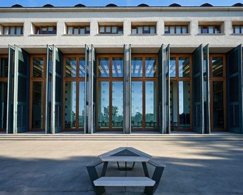 Ein modernes Gebäude mit großen Fenstern und offenen Fensterläden, die einen Blick auf den Innenraum ermöglichen. Vor dem Gebäude steht ein minimalistischer Tisch mit Bänken.