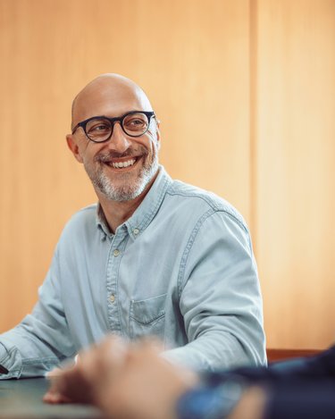 Ein lächelnder Mann mit Brille sitzt an einem Tisch und hält ein Tablet, während zwei weitere Personen im Hintergrund arbeiten.