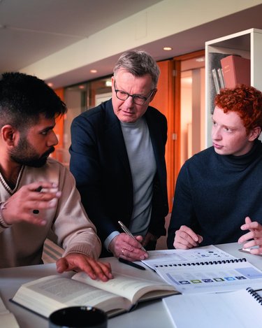 Drei Personen diskutieren an einem Tisch, während sie Notizen und Bücher verwenden. Ein Mann erklärt etwas, während die anderen aufmerksam zuhören.