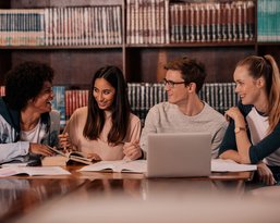Vier Studierende sitzen an einem Tisch in einer Bibliothek, umgeben von Büchern. Sie diskutieren und arbeiten an ihren Notizen und einem Laptop.