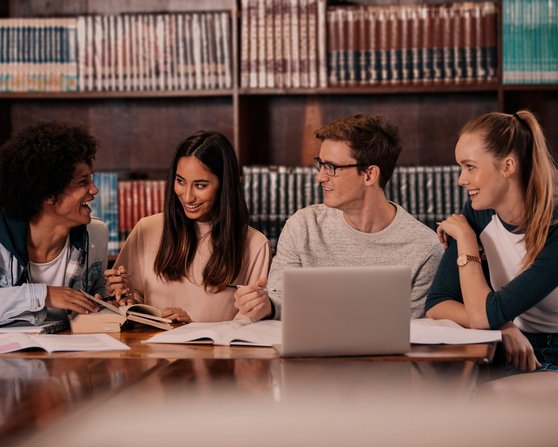 Vier Studierende sitzen an einem Tisch in einer Bibliothek, umgeben von Büchern. Sie diskutieren und arbeiten an ihren Notizen und einem Laptop.