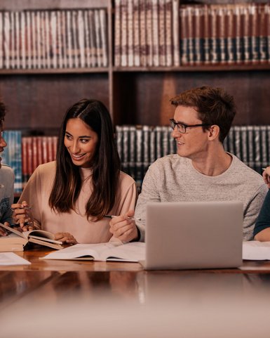 Four students are sitting at a table in a library, surrounded by books. They are discussing and working on their notes and a laptop.