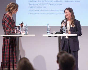 Zwei Frauen stehen an Stehtischen und sprechen miteinander, während Wasserflaschen und Gläser auf den Tischen stehen. Im Hintergrund sind Informationen zu sehen.