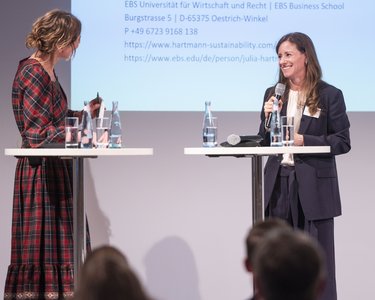 Two women are standing at high tables, chatting with each other, while water bottles and glasses are placed on the tables. In the background, information is visible.