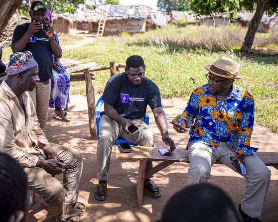 A group of men is sitting on benches under a tree, while two men in the middle are having a conversation.