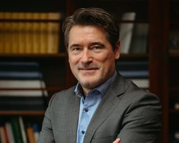 Ein Mann in einem grauen Anzug mit blauem Hemd steht lächelnd mit verschränkten Armen vor einem Bücherregal.