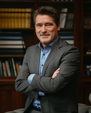 Ein Mann in einem grauen Anzug mit blauem Hemd steht lächelnd mit verschränkten Armen vor einem Bücherregal.