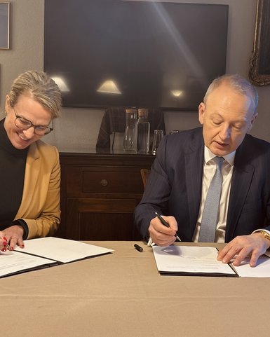 Zwei Personen unterzeichnen Dokumente an einem Tisch in einem Innenraum. Eine Frau trägt einen beigen Blazer, der Mann einen Anzug.