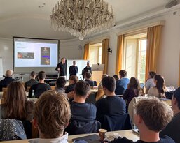 Eine Gruppe von Menschen sitzt in einem Seminarraum, während zwei Referenten eine Präsentation an einem Bildschirm halten.