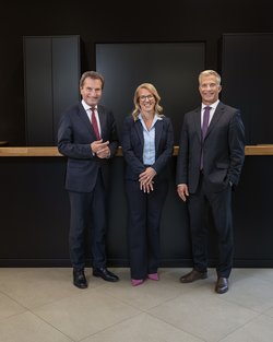 Three people in formal attire are standing side by side in a modern office building. The woman in the middle has short hair and is wearing glasses, while the two men on her left and right are smiling.