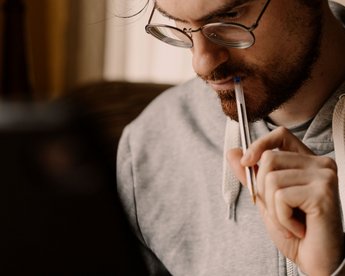 Ein Mann mit Brille hält einen Stift an seinen Lippen und schaut konzentriert auf einen Laptop.