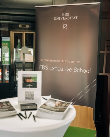 Roll-up-Banner der EBS Universität mit dem Slogan "Empowerment. Made by EBS." und Informationen zur EBS Executive School. Ein Tisch mit Broschüren und QR-Code ist sichtbar.